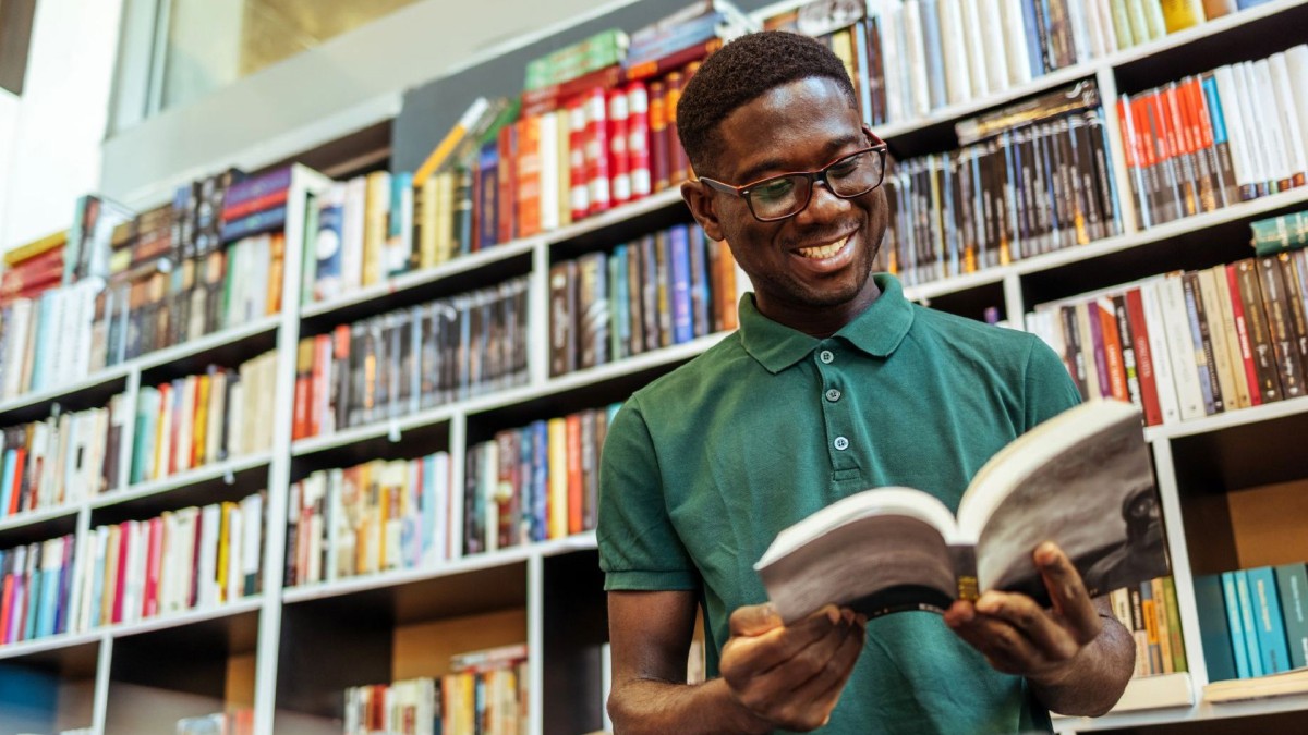 A man wearing glasses smiles as he reads a book in his school’s library.