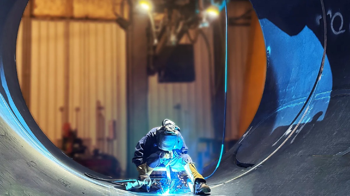 In an industrial workspace, a worker in protective gear is welding inside a large cylindrical metal structure as bright blue sparks fly.