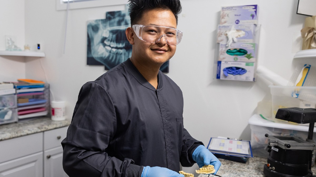 A man wearing safety glasses and a blue lab coat smiles as he holds a mold of teeth at a dental office.