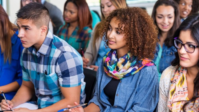 Students listen attentively while taking notes during a lecture on a college campus.