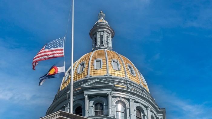 The U.S. and Colorado flags fly in front of a domed government building and blue sky.