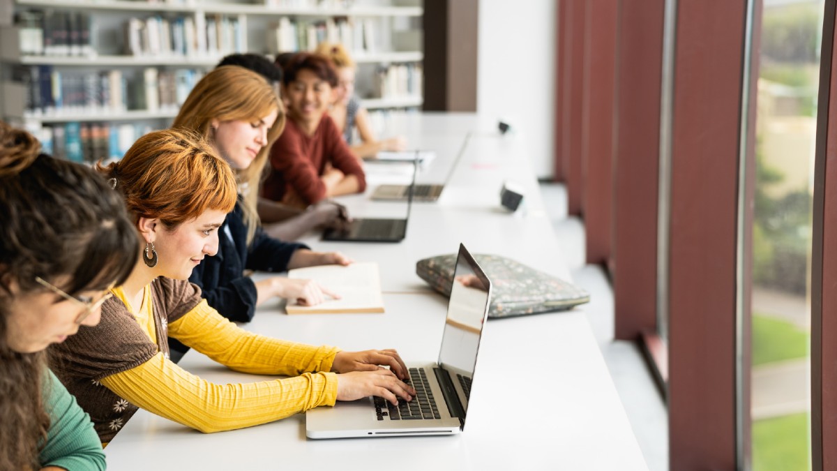 Several students are seen working on their laptops at a college campus library.