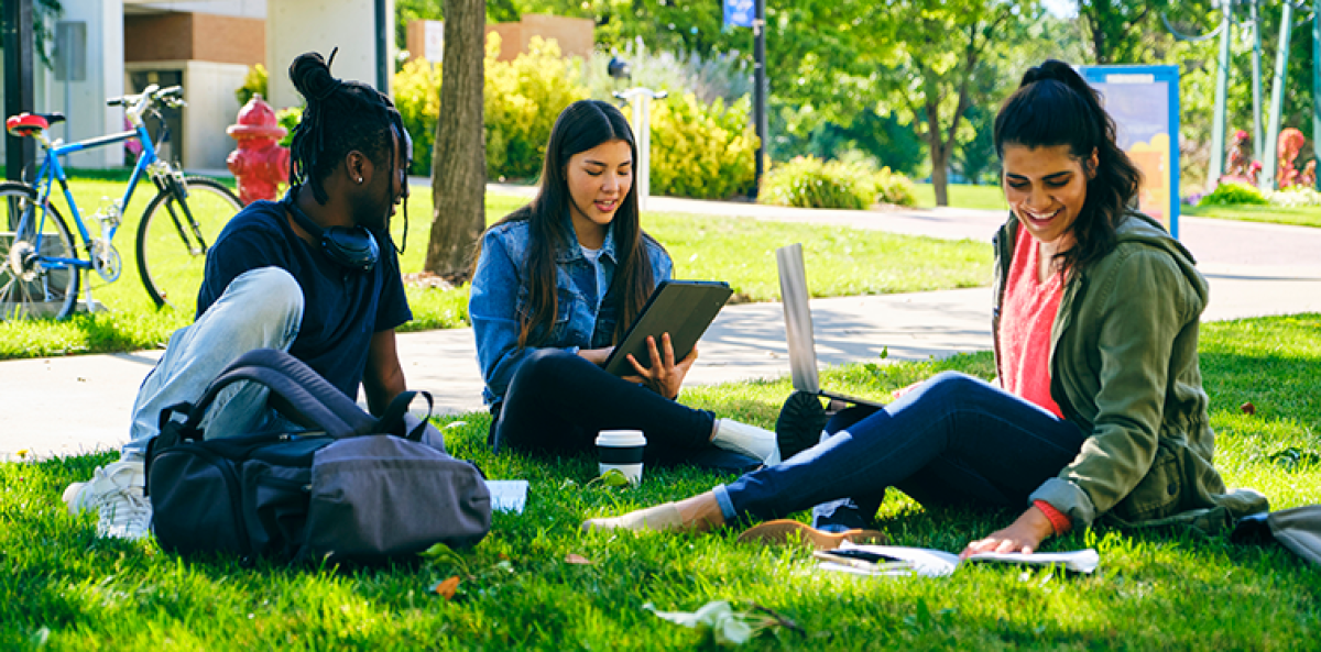 Three college students sit on grass in the quad at their institution, reviewing materials for an upcoming class.