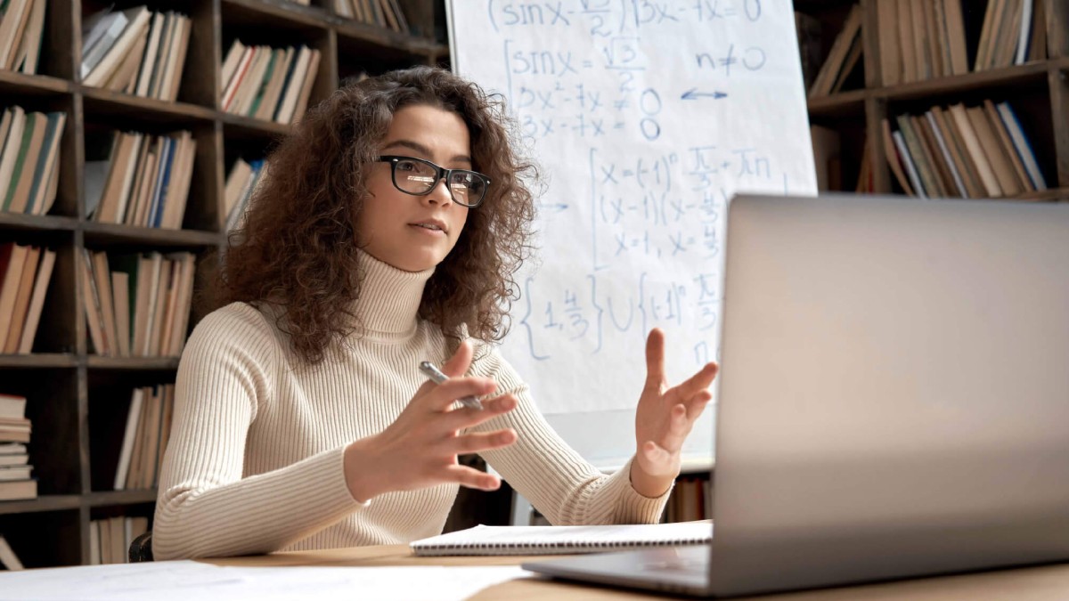 A curly haired woman is pictured in her office sharing insights during a video call on her laptop.
