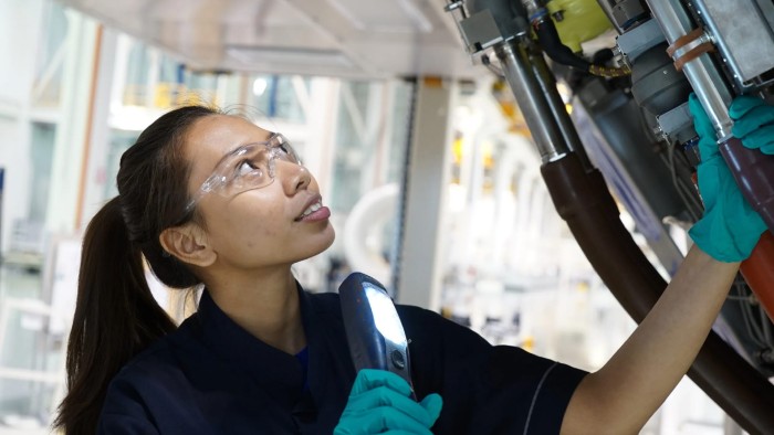 A woman holding a flashlight and wearing safety glasses inspects a mechanical component on a piece of machinery.