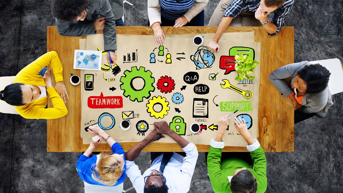 Eight people are seated around a wood table with a large sheet of paper at the center decorated with graphics that depict teamwork, communication, and critical thinking.