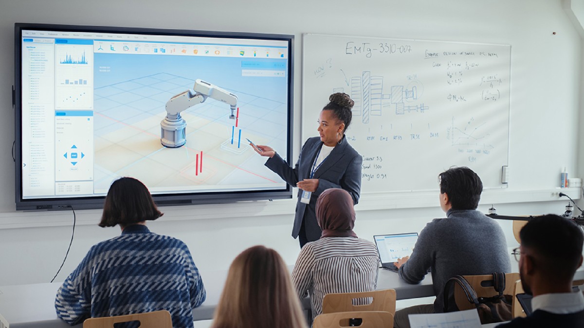 In a college classroom, a faculty member wearing a gray dress suit points to a large screen featuring a robot arm. Several students take notes while looking at the screen. A white dry-erase board to the right of the screen shows physics equations and diag