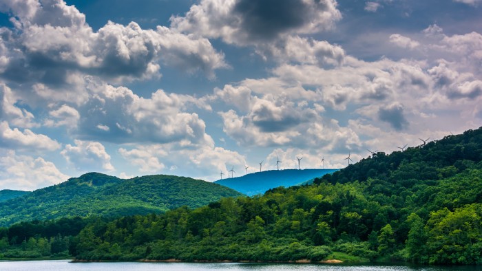 A blue sky with fluffy white clouds gives way to lush green rolling hills and a body of water, with wind turbines rotating in the distance.