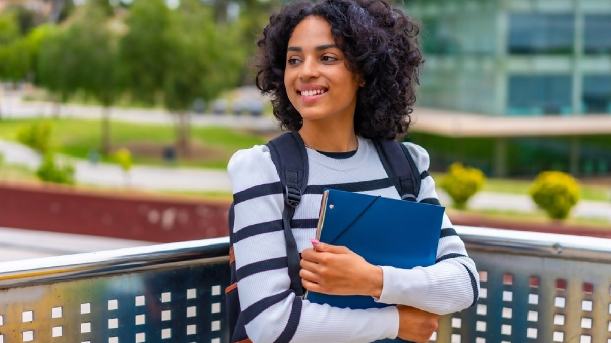 A woman with curly black hair smiles while clutching a notebook and wearing a backpack on a college campus.