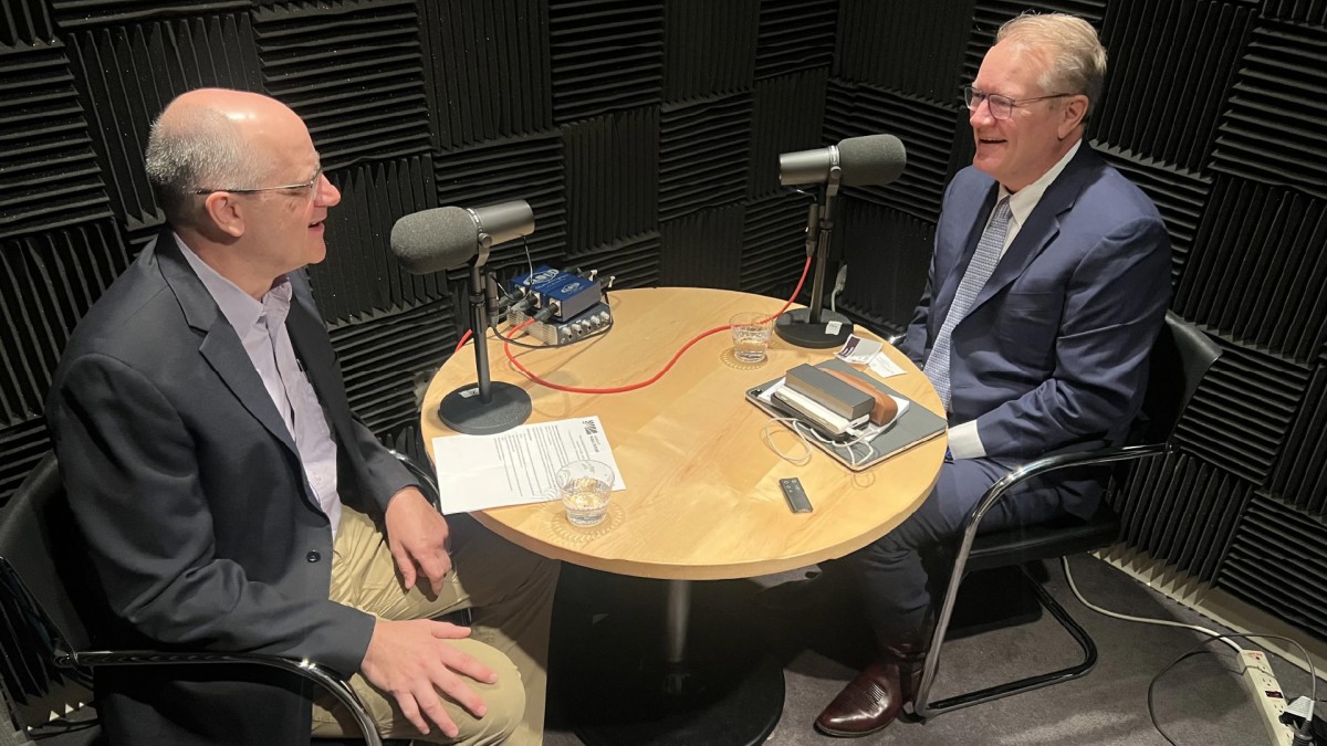 A podcast host and his interviewee sit at a table with microphones, notes, and glasses of water.