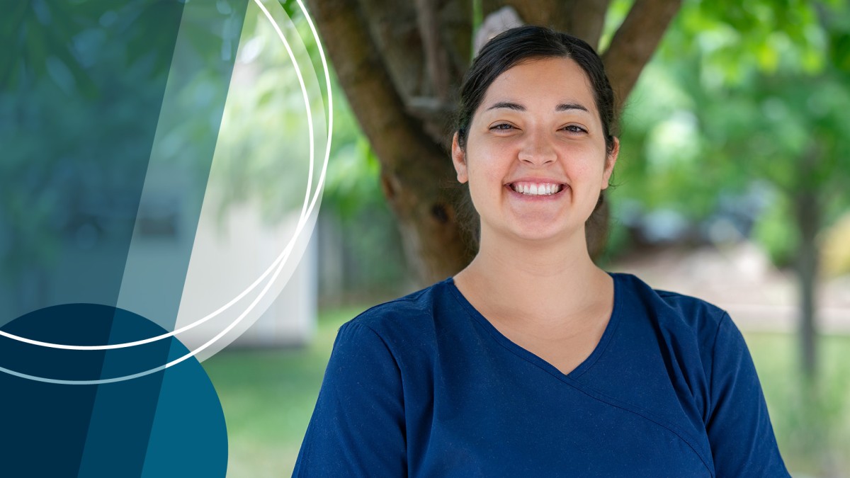 A smiling young woman in dark blue scrubs stands before a tree in a verdant courtyard. 