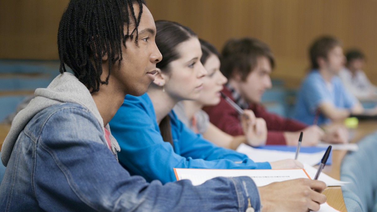 Students are pictured sitting at a table, listening closely while taking notes with pen and paper.