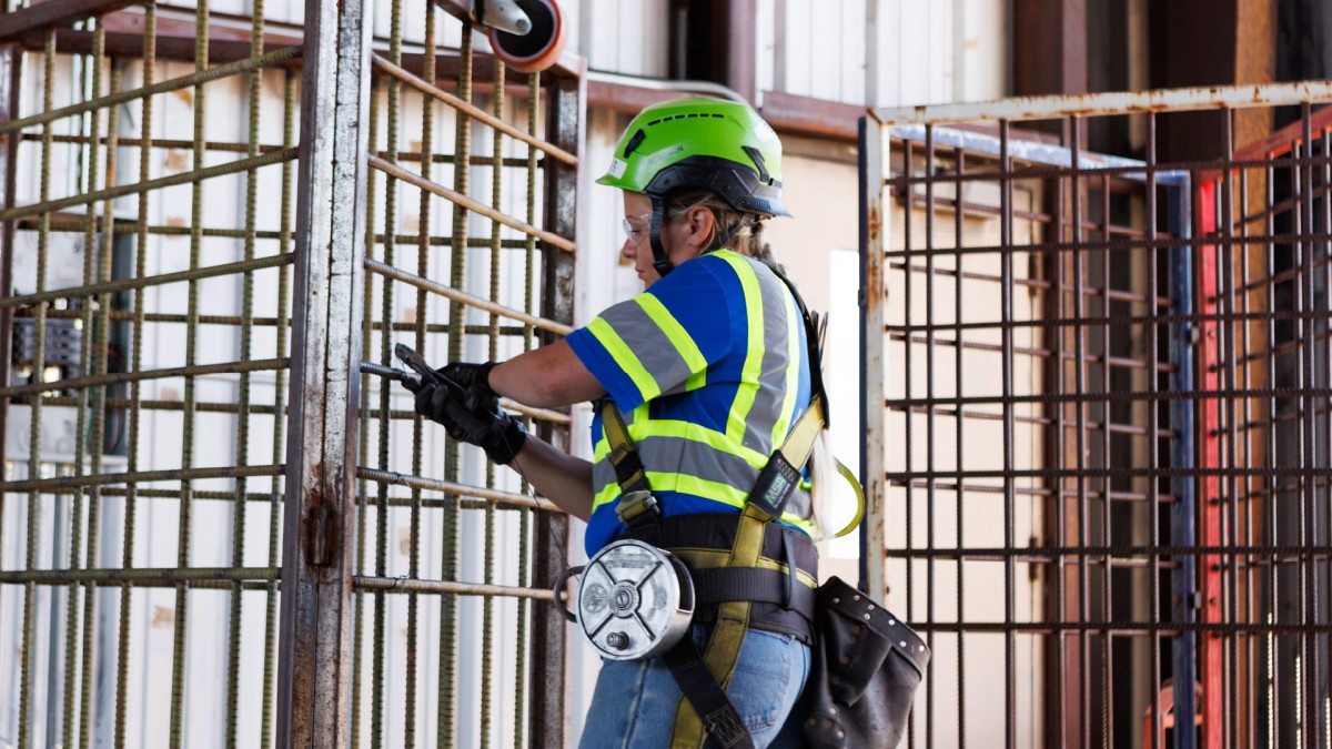 A woman wearing a hard hat and safety vest works on a metal structure as part of job training within a correctional facility.