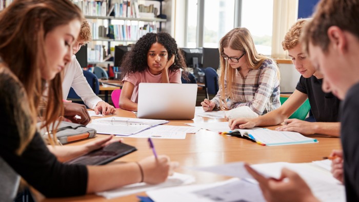 Students gather around a table, exchanging papers as they prepare for an English exam.