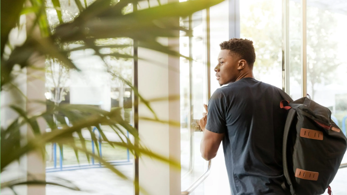 A male college student with a backpack opens a glass door to exit a building, with green plants in the foreground and natural light streaming in from outside.