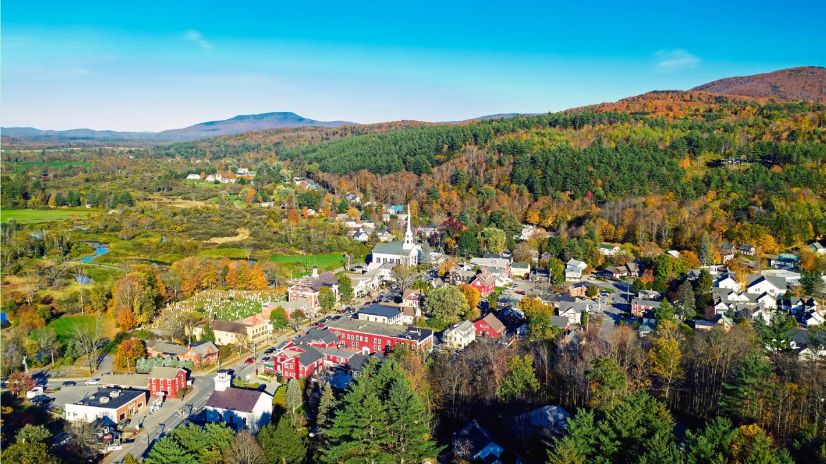 Aerial view of a rural town surrounded by forests and rolling hills in autumn. Colorful foliage blankets the landscape, and a white church with a steeple stands at the center of the town. Scattered houses, local businesses, and a cemetery are visible, wit