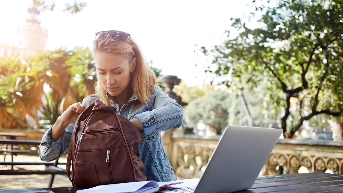 A young woman searches through her backpack while using a laptop and notebook to study for an exam outdoors.