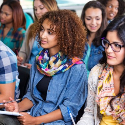 Students listen attentively while taking notes during a lecture on a college campus.