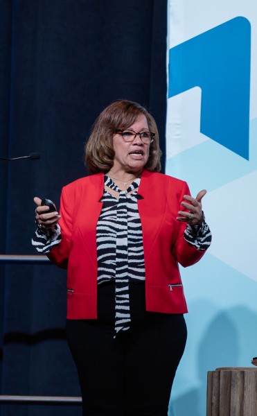 A woman in a red blazer speaks on stage to an audience