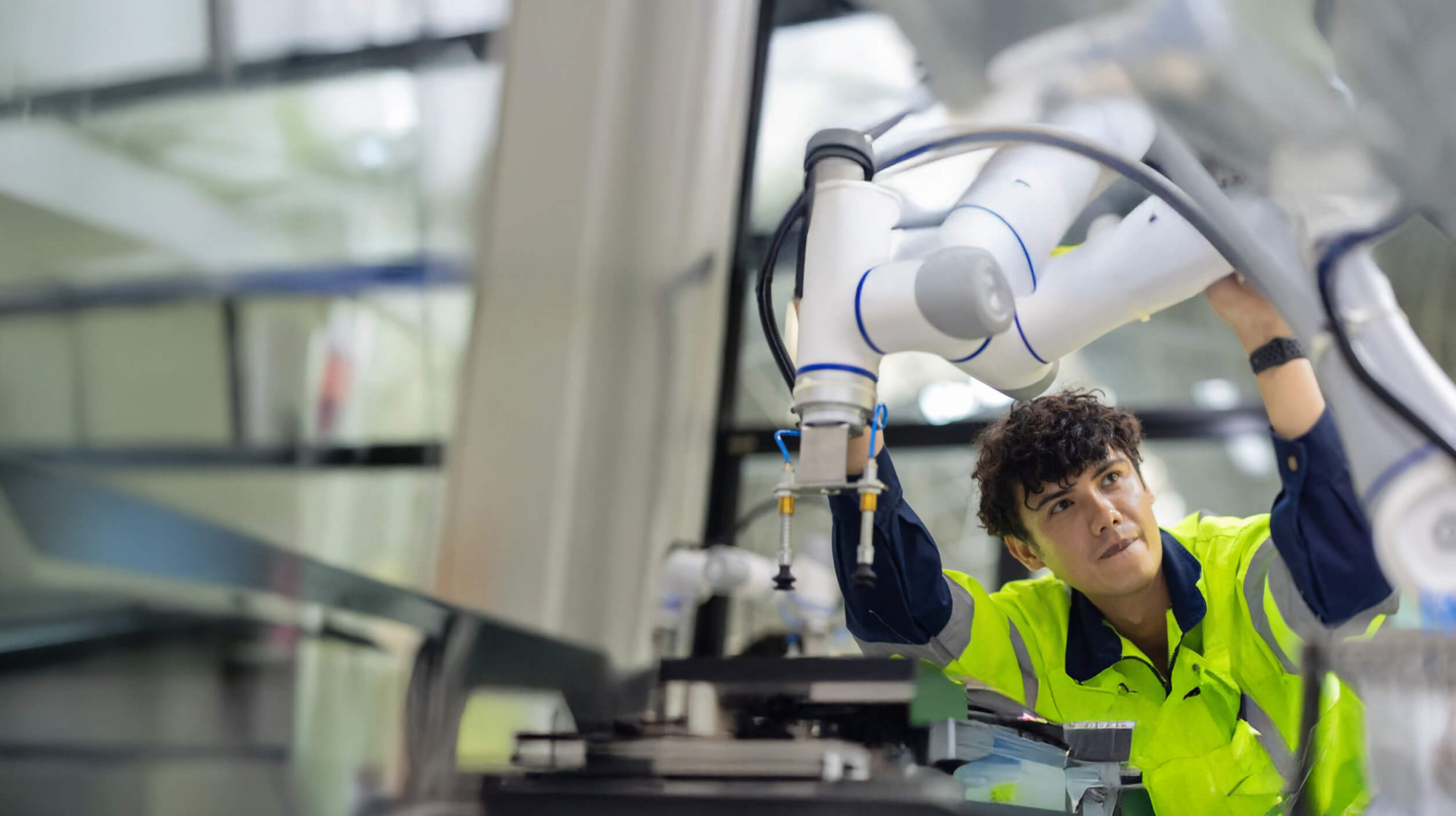 A young man in a fluorescent safety vest makes adjustments to a large white robotic arm