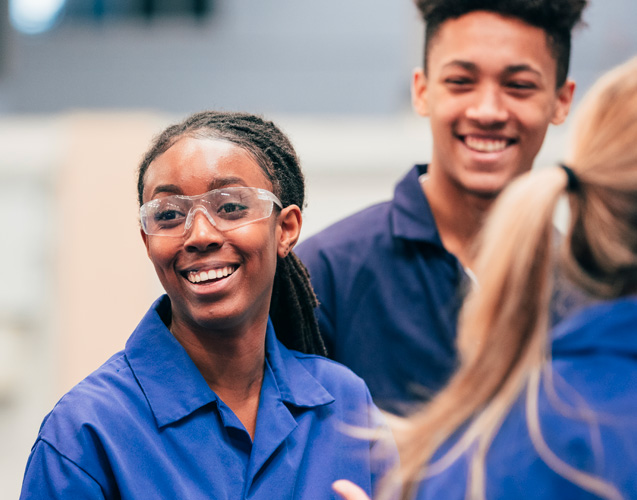 Smiling African American woman in blue working shirt surrounded by 2 colleagues. She is wearing safety glasses. 